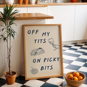 May include: A framed sign with the text "OFF MY TITS ON PICKY BITS" in navy blue. The sign has illustrations of a wine glass, cheese board, and a bowl of olives. A small potted plant and a basket of oranges are in the foreground.
