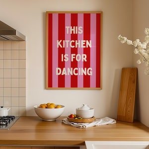May include: A framed print with a red and pink striped design and the words "THIS KITCHEN IS FOR DANCING". The print is displayed in a kitchen above a wooden work surface with a bowl of fruit and other kitchen items.