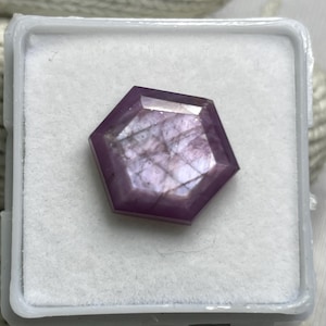 Pode incluir: Uma pedra preciosa hexagonal facetada com um tom roxo e rosa translúcido, apresentando inclusões internas e uma superfície polida. A pedra preciosa está colocada com segurança dentro de uma caixa de exibição quadrada branca.
