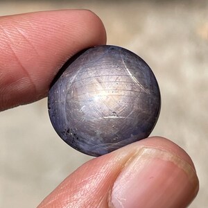 Pode incluir: Uma pedra preciosa redonda e polida, segurada entre dois dedos. A gema é de uma cor azul-acinzentada escura com um padrão em forma de estrela em sua superfície. A pedra tem uma aparência lisa e reflexiva, com variações sutis de cor e textura.