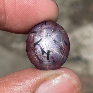 May include: A round, polished gemstone with a star effect. The gem is held between two fingers. The stone has a deep purple hue with black inclusions and a six-rayed star pattern across its surface.