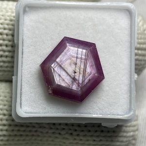 Op de afbeelding: Een vijfhoekige, diep magenta gekleurde edelsteen met zichtbare interne lineaire insluitingen en een gefacetteerd oppervlak, gepresenteerd in een kleine, vierkante, witte plastic houder met een witte schuimrubberen inzet.