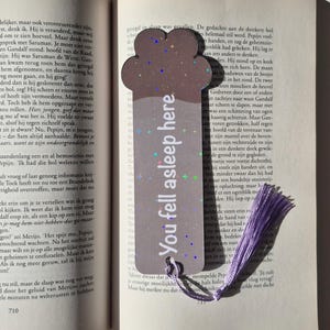 May include: A brown bookmark with a cloud-like top and a purple tassel. The text "You fell asleep here" is printed on the bookmark with a holographic effect. The bookmark is placed in an open book.