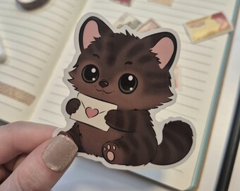 May include: A sticker featuring a cartoon kitten holding an envelope with a heart. The kitten is brown with black stripes and large, expressive eyes. The envelope is white with a pink heart. The sticker is on a notebook.