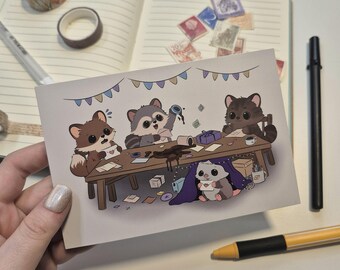 May include: A postcard featuring an illustration of four raccoons and a hedgehog around a table. The raccoons are depicted with various expressions, and the table is decorated with small gifts, coffee cups, and spilled ink. A hand holds the postcard.