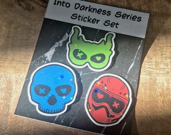 May include: A sticker set titled Into Darkness Series Sticker Set featuring three holographic stickers on a black marble-patterned backing card. The stickers include a green flame-shaped mask, a blue skull, and a red helmet with goggles.