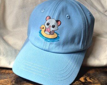 May include: Light blue baseball cap featuring a cartoon hamster floating on a yellow duck-shaped inflatable ring. The hamster has large eyes and is holding a seashell. The cap has a curved brim and an adjustable strap.