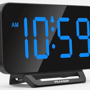 May include: A black digital alarm clock with a rectangular display featuring bright blue LED numbers showing 10:59 AM. The device has a sleek, rounded-edge screen and a sturdy black base with the brand name PEAKEEP printed in white on the front.