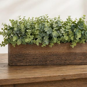 May include: A rectangular wooden planter box filled with faux eucalyptus greenery. The box has a dark brown finish and sits on a wooden table. Decorative home decor.