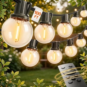 May include: String lights with clear, round bulbs and black fixtures are strung across a green background. A tag reads "100 FT". A remote control is in the foreground. The lights are a warm yellow color.