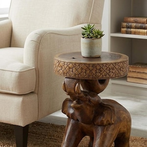 May include: A brown wooden side table featuring a carved elephant base and a circular top with a floral pattern along the edge. A small, light-colored ceramic pot containing a green succulent plant sits on the flat surface of the table.