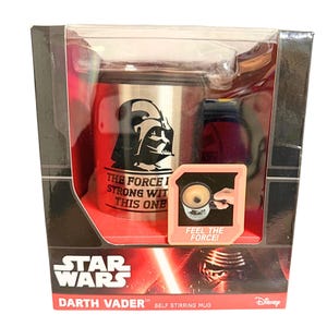 May include: A silver-toned stainless steel self-stirring mug featuring a black graphic of Darth Vader and the text THE FORCE IS STRONG WITH THIS ONE. The packaging includes a Star Wars logo and a small inset image with the text FEEL THE FORCE.