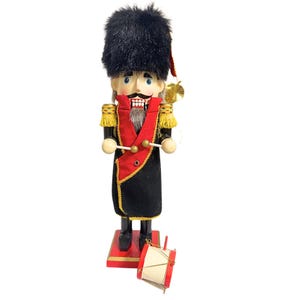 May include: A wooden nutcracker soldier in a black uniform with gold trim, a red sash, and a tall black furry hat. The nutcracker holds drumsticks and stands on a red base. A small red and white drum sits at its feet.