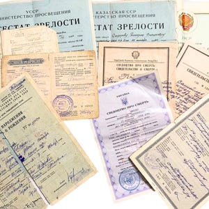 Op de afbeelding: Een verzameling vintage documenten, waaronder certificaten en officiële papieren, met tekst in het Cyrillisch schrift. De papieren zijn verouderd met zichtbare slijtage, stempels en handgeschreven aantekeningen. De documenten zijn in verschillende formaten en kleuren.