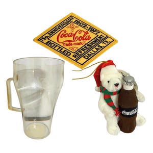 May include: A clear glass Coca-Cola mug with a handle, a yellow patch with "75th Anniversary Coca-Cola" text, and a polar bear ornament holding a Coca-Cola bottle. The patch has the text "Bottled Refreshment Dallas, TX."