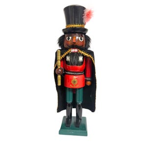 Puede incluir: Figura de cascanueces de madera, vestida con un sombrero de copa negro con una pluma rosa, una chaqueta roja, pantalones verdes y una capa negra. La figura sostiene un bastón dorado y se encuentra sobre una base verde. Mide unos 30 cm de alto.