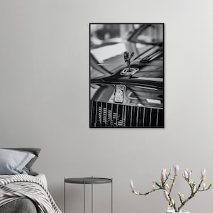 May include: Black and white framed print of a luxury car. The image focuses on the front of the car, showcasing the hood ornament, the Rolls-Royce emblem, and the grille. The print is displayed on a white wall.