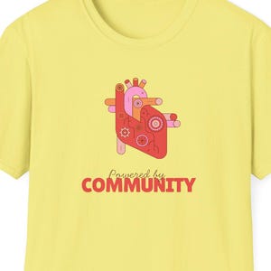 May include: A light yellow t-shirt featuring a stylized anatomical heart design in pink, red, and orange. The text "Powered by COMMUNITY" is printed below the heart in red. The shirt has short sleeves and a crew neck.