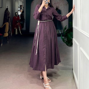 Puede incluir: Un vestido largo abotonado de color ciruela oscuro. El vestido tiene mangas largas, cuello camisero y falda plisada. Un cinturón de cuerda blanca ciñe la cintura. La persona lleva sandalias blancas con tacón cuadrado.
