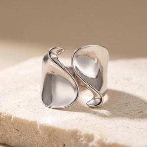 Peut inclure: Une bague en métal argenté poli présentant un design ouvert avec deux éléments lisses, incurvés et en forme de pétales qui s'enroulent autour du doigt. La surface métallique est très réfléchissante, créant une forme fluide et organique.
