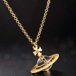 Peut inclure: Un collier doré avec un pendentif en forme de planète entouré d'un anneau. Le pendentif comprend une sphère transparente, une croix surmontée d'une petite pierre claire et des pierres colorées incrustées le long de la structure de l'anneau.