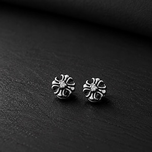 Può includere: Un paio di orecchini a bottone color argento. Ogni orecchino presenta un design floreale con una piccola gemma trasparente al centro. Gli orecchini sono esposti su una superficie scura e strutturata.