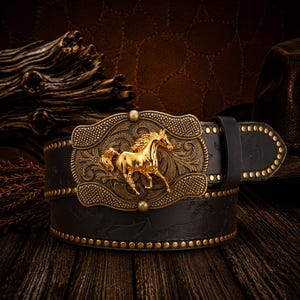 May include: A black leather belt featuring a large, rectangular gold-tone buckle with an ornate, embossed floral pattern and a prominent, three-dimensional gold horse figure in the center. The belt strap is decorated with small gold studs.