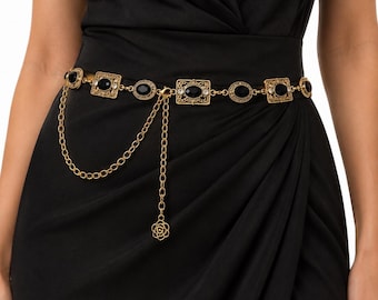 May include: A gold-tone chain belt featuring alternating rectangular and oval filigree links set with black stones. The belt includes a dangling chain section with a small flower-shaped charm, worn over a solid black wrap-style dress.