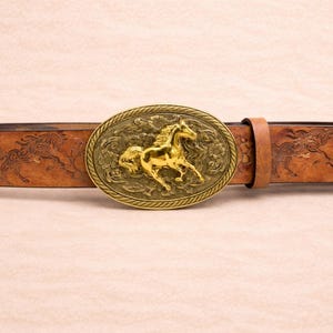 May include: A brown leather belt with an oval, gold-colored buckle featuring a running horse. The belt has an embossed design of horses and floral patterns. The buckle is approximately 3 inches wide.