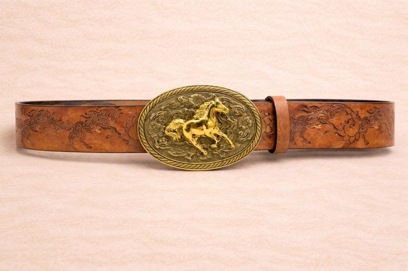 May include: A brown leather belt with an oval, gold-colored buckle featuring a running horse. The belt has an embossed design of horses and floral patterns. The buckle is approximately 3 inches wide.