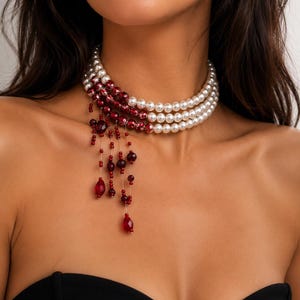 Peut inclure: Un collier ras-du-cou multi-rangs composé de trois rangées de perles blanches qui se transforment en perles rouge foncé sur un côté. Plusieurs fils délicats de perles rouges et de cristaux en forme de goutte pendent sur le côté du collier.
