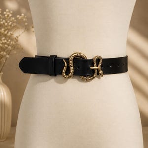 Pode incluir: Um cinto preto com uma fivela metálica em tom dourado em forma de serpente enrolada. O cinto tem um acabamento liso e uma extremidade pontiaguda, com um passador preto a condizer para prender a correia no lugar à volta da cintura.