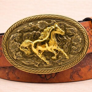 May include: A brown leather belt with a large, gold-colored oval buckle. The buckle features a detailed relief of a running horse, surrounded by floral designs. The belt itself has an embossed horse pattern.