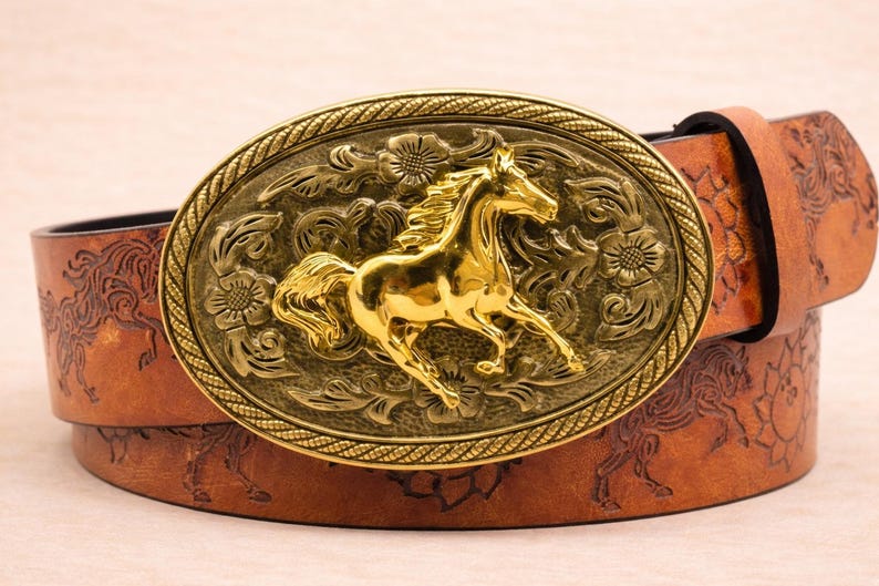 May include: A brown leather belt with a large, gold-colored oval buckle. The buckle features a detailed relief of a running horse, surrounded by floral designs. The belt itself has an embossed horse pattern.