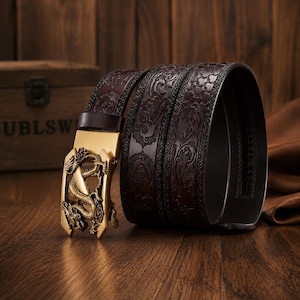 Op de afbeelding: Een donkerbruine leren riem met een ingewikkeld reliëfpatroon over de band en een goudkleurige metalen gesp in de vorm van een draak. Het gespmechanisme is rechthoekig met een gedetailleerd uitgesneden ontwerp van een draak.