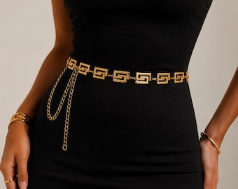 May include: A gold-toned metal chain belt featuring a repeating rectangular Greek key pattern. The belt includes a dangling chain section that hangs from the waistline, providing an adjustable length. It is worn over a plain black strapless dress.