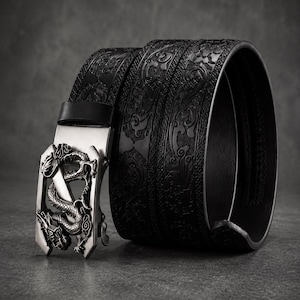 Puede incluir: Un cinturón negro con un patrón en relieve en toda la correa y una hebilla de metal plateado con forma de dragón. La hebilla presenta un diseño de dragón tridimensional detallado con escamas y garras intrincadas, mientras que la correa muestra un motivo decorativo repetitivo.