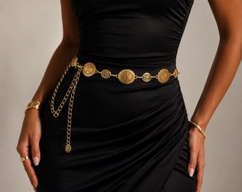 Pode incluir: Um cinto de corrente em tom dourado com medalhões circulares com detalhes em relevo, usado sobre um vestido preto sem alças com franzido lateral e uma fenda alta na perna. O cinto inclui uma seção de corrente pendente com um pequeno pingente floral.