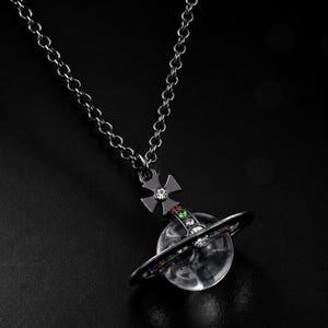 Peut inclure: Un collier à chaîne en métal sombre avec un pendentif en forme de planète entouré d'un anneau. Le pendentif comprend un centre sphérique transparent, une pièce supérieure en forme de croix avec une pierre claire et des cristaux colorés.