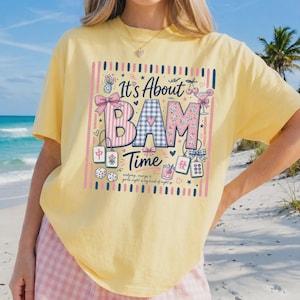 May include: A pale yellow short-sleeve t-shirt featuring a graphic print with the text It's About BAM Time. The design includes illustrations of mahjong tiles, dice, cherries, a cocktail glass, and bows, framed by vertical pink and blue stripes.