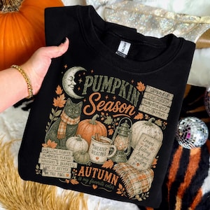 May include: A black sweatshirt featuring a graphic print with a crescent moon, a cat wearing a scarf, pumpkins, a lantern, and a mug labeled Sweater Weather. Text includes Pumpkin Season, Autumn is my favorite color, and fall phrases.