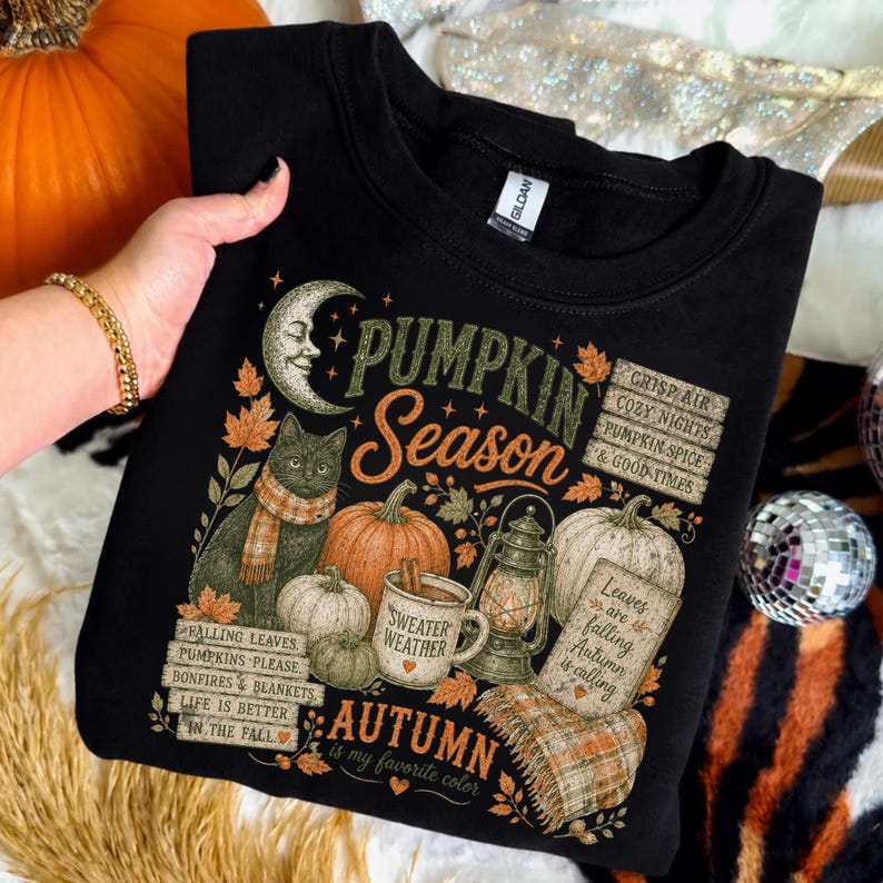 May include: A black sweatshirt featuring a graphic print with a crescent moon, a cat wearing a scarf, pumpkins, a lantern, and a mug labeled Sweater Weather. Text includes Pumpkin Season, Autumn is my favorite color, and fall phrases.