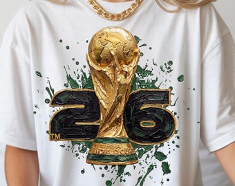 Może przedstawiać: Biała koszulka z grafiką złotego trofeum FIFA World Cup i numerem "26" w kolorze czarnym i zielonym. Projekt zawiera zielone plamy farby. Koszulka ma okrągły dekolt i krótkie rękawy.