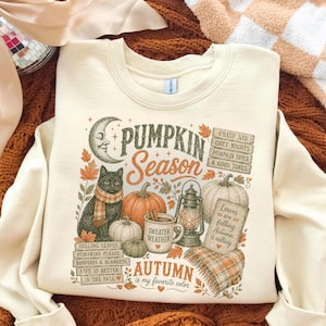 May include: A cream-colored crewneck sweatshirt featuring a graphic print with a black cat, pumpkins, a lantern, and a crescent moon. The text reads: PUMPKIN SEASON, CRISP AIR COZY NIGHTS PUMPKIN SPICE & GOOD TIMES, SWEATER WEATHER, AUTUMN is my favorite color.