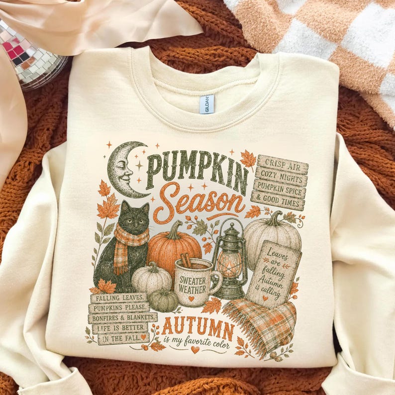 May include: A cream-colored crewneck sweatshirt featuring a graphic print with a black cat, pumpkins, a lantern, and a crescent moon. The text reads: PUMPKIN SEASON, CRISP AIR COZY NIGHTS PUMPKIN SPICE & GOOD TIMES, SWEATER WEATHER, AUTUMN is my favorite color.