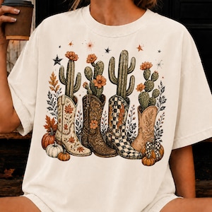 Puede incluir: Una camiseta color hueso con un estampado gráfico de cuatro botas vaqueras, cada una con un cactus verde y flores naranjas. El diseño incluye hojas de otoño, calabazas y pequeñas estrellas. Las botas presentan patrones florales, lisos y a cuadros.
