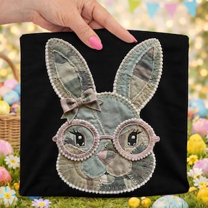May include: Black tote bag featuring a patchwork bunny face with glasses and a bow. The bunny's face is made of various fabrics in shades of green, gray, and pink, with pearl-like trim. The glasses are pink, and the bow is a matching pink.