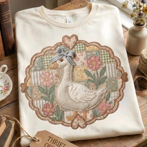 May include: Cream-colored t-shirt featuring a detailed appliqué of a goose with a blue bow, surrounded by a patchwork design of floral and gingham patterns. The design includes pink flowers and heart accents, creating a vintage, cottagecore aesthetic.