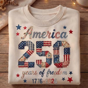 May include: Cream-colored sweatshirt with patriotic embroidery. Features the word "America" and the number "250" in a patchwork design, along with the phrase "years of freedom" and the dates 1776-2026. Red and blue stars complete the design.