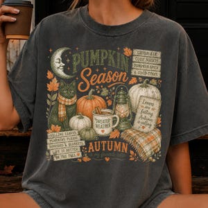 May include: A charcoal gray t-shirt featuring a graphic print with a crescent moon, a cat in a scarf, pumpkins, a lantern, and a mug labeled "Sweater Weather." The text includes "Pumpkin Season," "Autumn," and various phrases about fall.